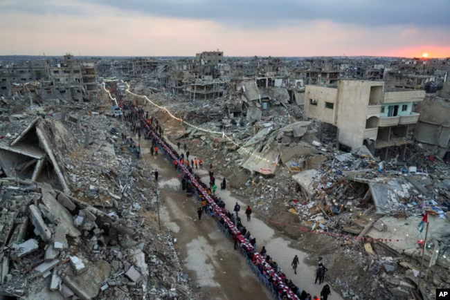 Saat matahari terbenam, warga Palestina duduk di meja besar yang dikelilingi puing-puing rumah dan bangunan ya